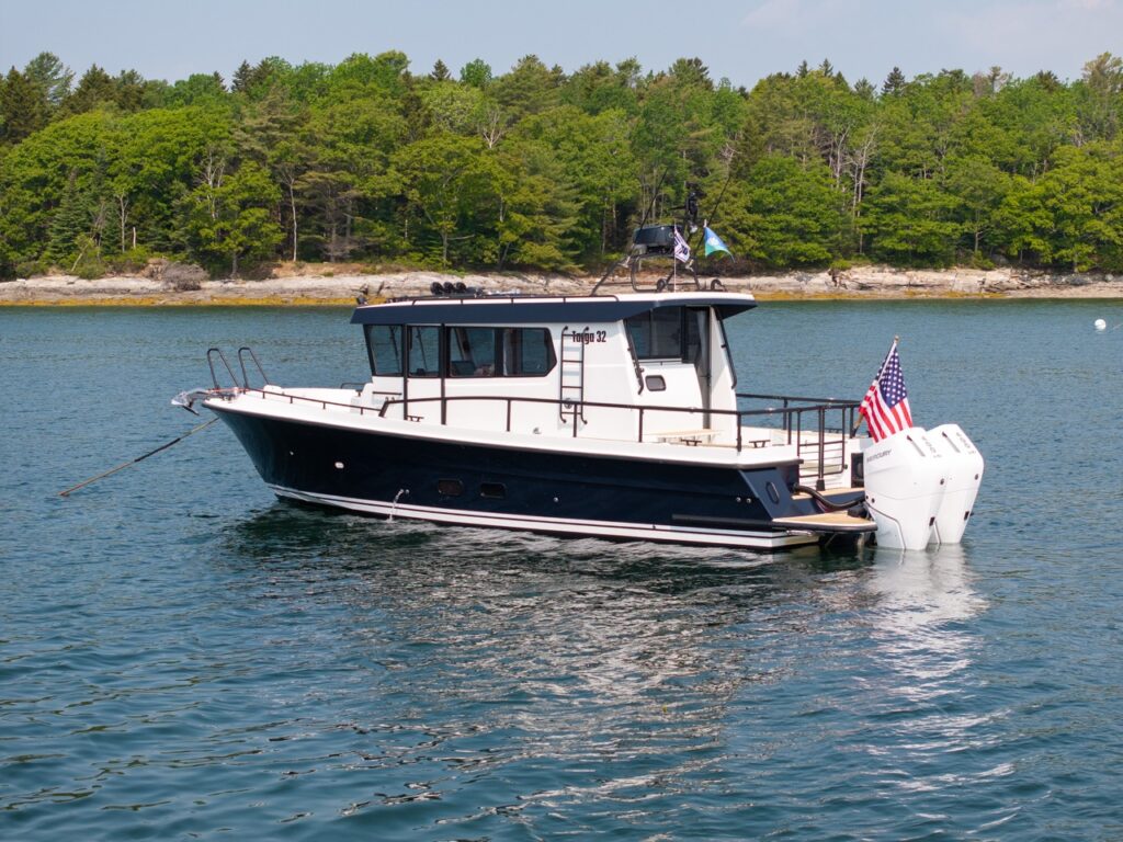 Starboard-side profile of a Targa 32 pilothouse boat at anchor on calm coastal water, featuring an enclosed cabin, walkaround decks, aft cockpit, and twin Mercury Verado outboards flying an American flag.