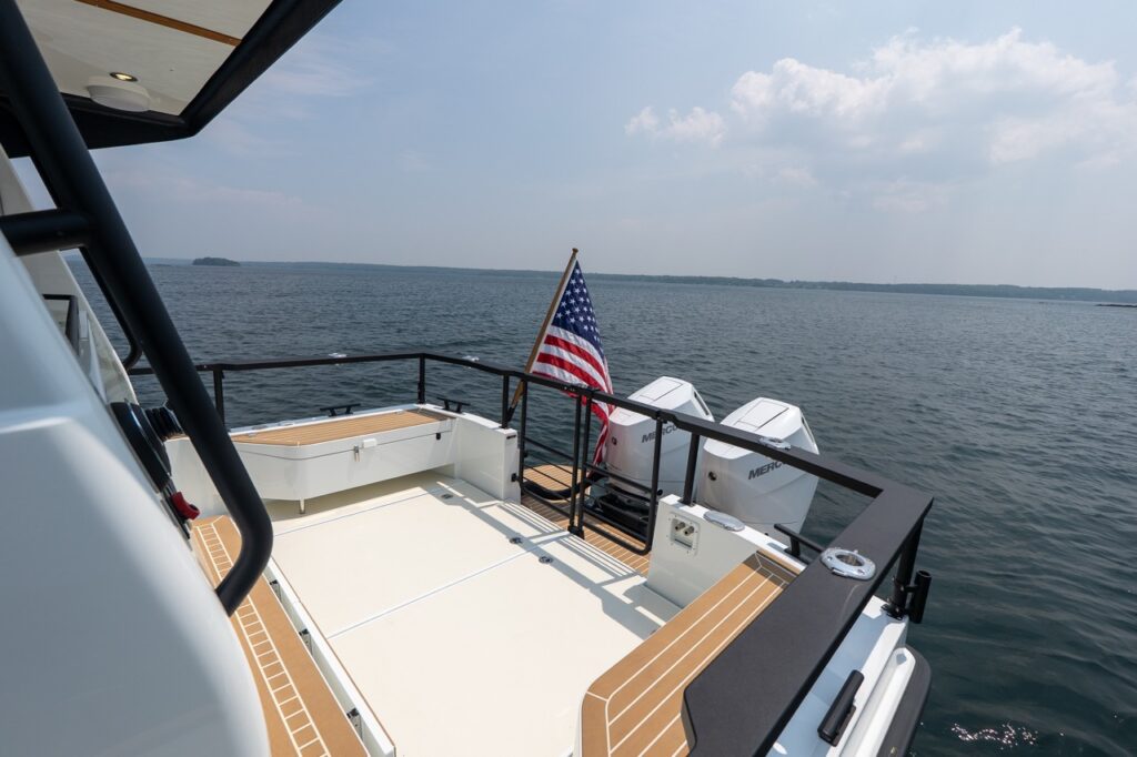 Wide view of the Targa 32 aft cockpit with open deck space, teak-style decking, black safety rails, an American flag at the stern, and twin Mercury Verado outboards beyond.