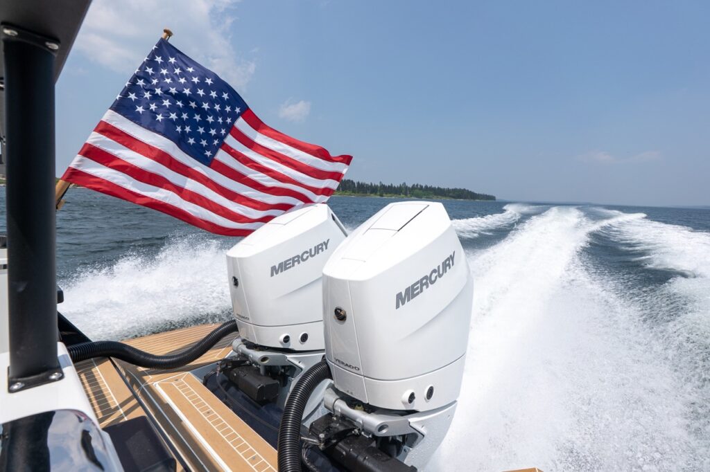 Twin white Mercury Verado outboard engines powering a Targa 32 at cruising speed, with an American flag flying above and a wide wake trailing behind on open water.