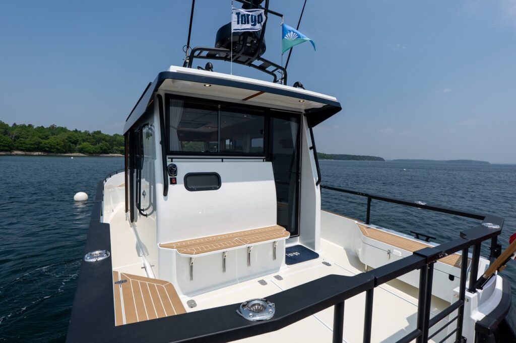Aft cockpit of a Targa 32 with a centered transom door, teak decking, fold-down transom bench seating, black safety rails, and the pilothouse aft window, set against calm coastal waters.