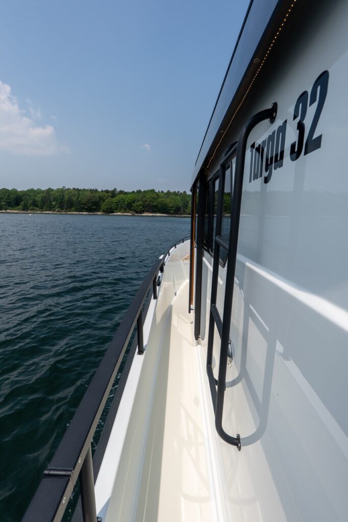 Port side deck walkway on a Targa 32, featuring a wide, protected side deck with tall black handrails, pilothouse grab rails, and calm coastal water alongside a wooded shoreline.