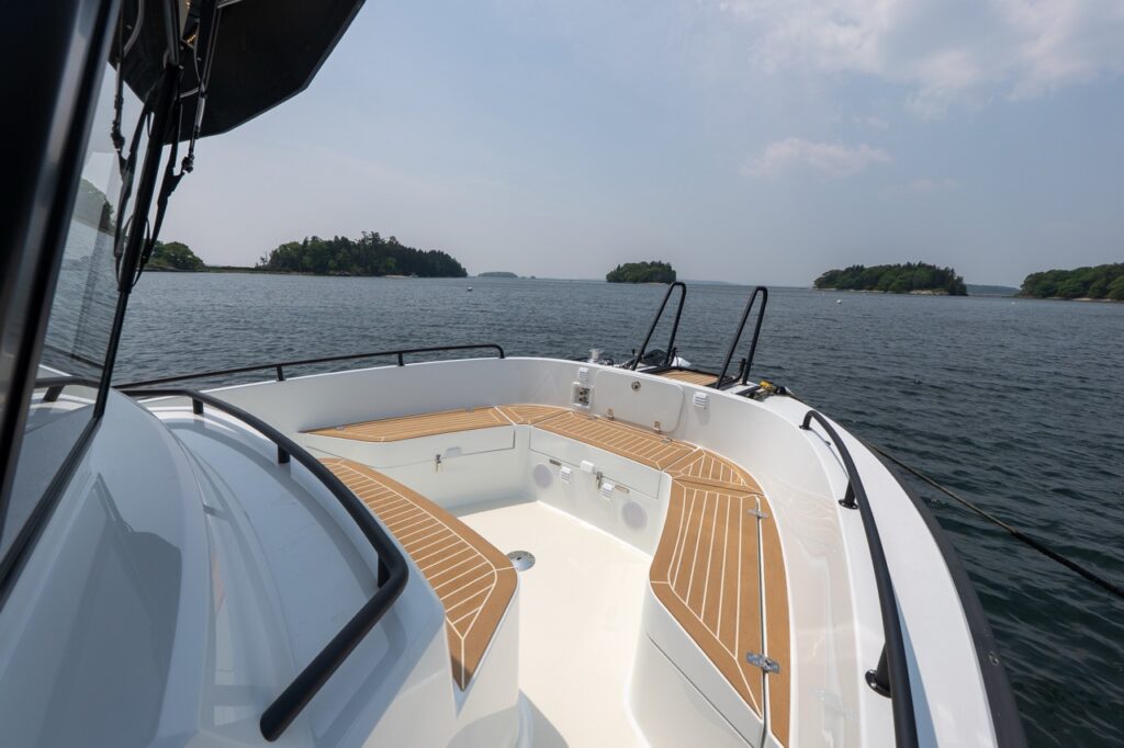 Forward bow seating area on the Targa 32 with wraparound benches, teak decking, integrated speakers, and black safety rails, viewed from the port side with open water and islands ahead.