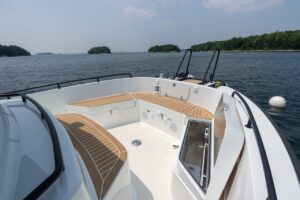 Open bow seating area on the Targa 32 featuring wraparound benches with teak decking, integrated speakers, stainless storage access on the starboard side, and black safety rails overlooking calm coastal waters.
