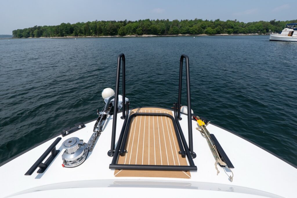 Forward foredeck of the Targa 32 with teak decking, black safety rails, anchor windlass, and bow hardware, overlooking calm water and a wooded shoreline ahead.