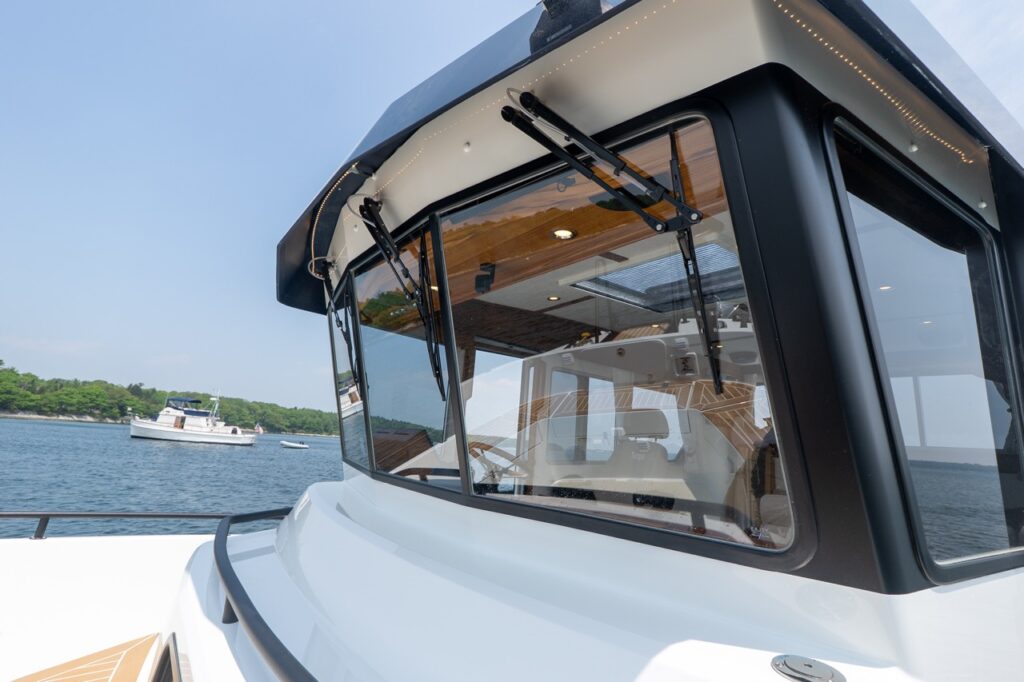 Exterior view of the Targa 32 pilothouse showing large wraparound windows with dual windshield wipers, black window frames, and teak foredeck below, with calm coastal waters in the background.