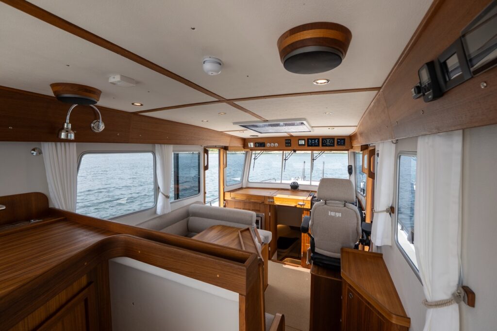 Interior view of the Targa 32 pilothouse looking forward, showing the port-side dinette, starboard helm seat, teak cabinetry, overhead lighting, and large windows with water visible outside.
