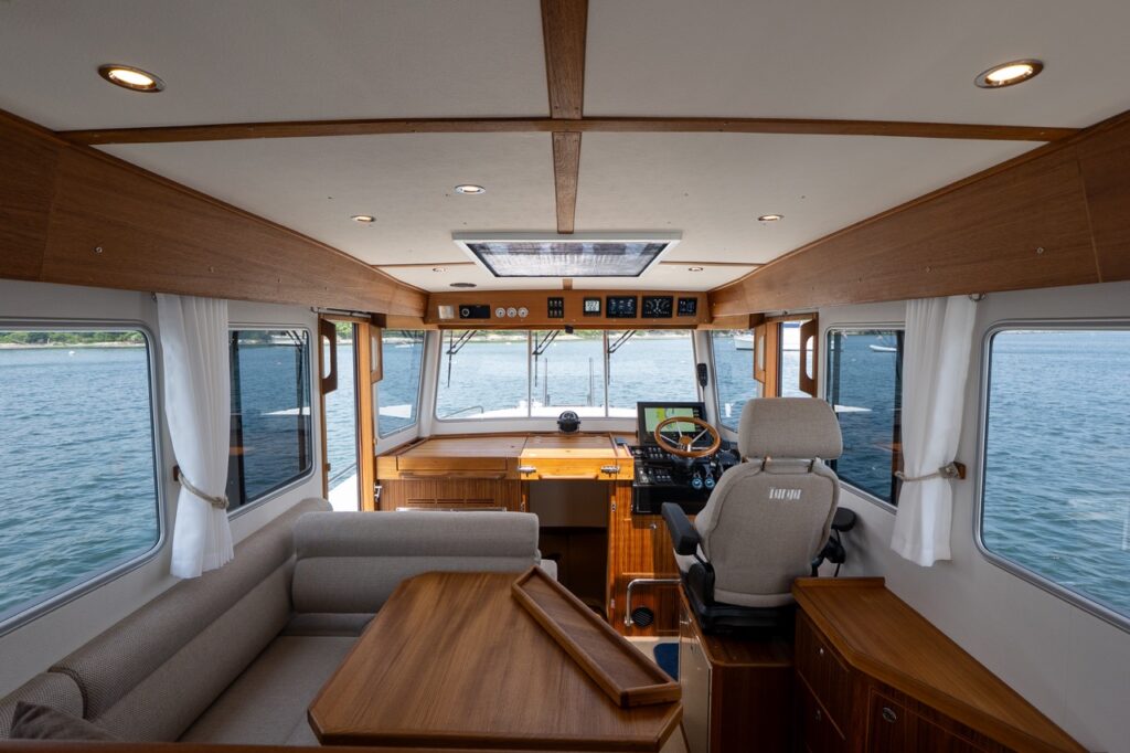 Wide view of the Targa 32 pilothouse interior showing the port-side dinette, teak table, helm station to starboard, panoramic windows, and overhead skylight with ocean visible outside.
