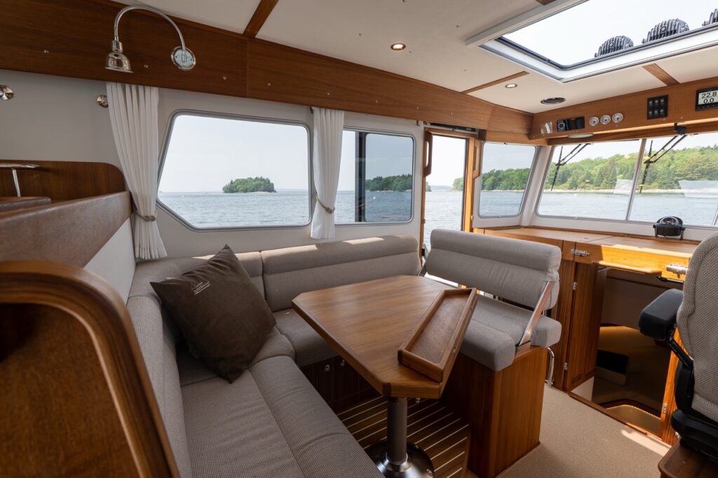 Targa 32 pilothouse interior showing the port-side dinette with upholstered seating, teak table, large windows, and forward helm area with natural light.