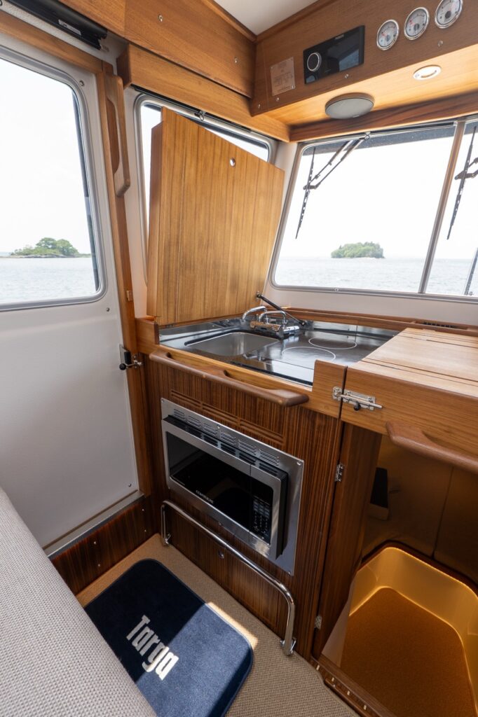 Pilothouse galley on a Targa 32 with teak cabinetry, lift-up countertop lid, stainless sink, electric cooktop, and built-in microwave below.