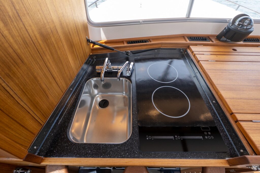 Compact galley on a Targa 32 with a stainless-steel sink and two-burner induction cooktop set into a black countertop, surrounded by teak cabinetry.