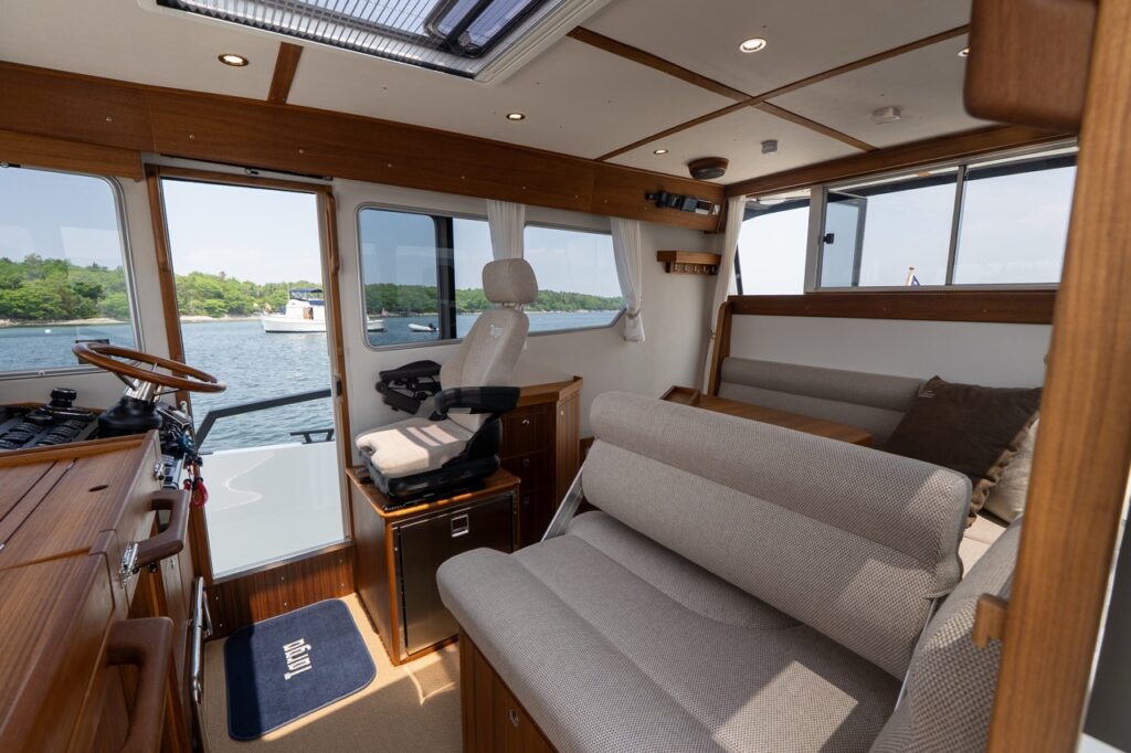 Targa 32 pilothouse interior showing the helm seat beside a sliding side door, a forward-facing settee with table, teak cabinetry, and panoramic windows overlooking the water.