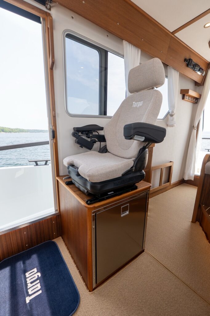 Targa 32 pilothouse helm seat positioned beside a sliding side door, with large windows, light upholstery, teak cabinetry, and a compact refrigerator below the seat.