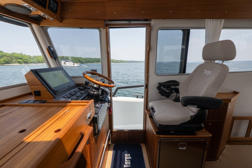 Pilothouse helm area with a wooden steering wheel, Multi Function Display, teak cabinetry, and a Targa helm seat beside an open sliding door leading to the starboard side deck.