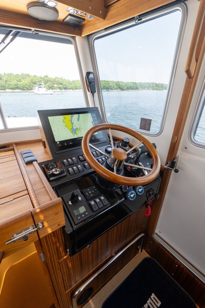 Starboard-side helm station with a wooden steering wheel, teak cabinetry, and a Multi Function Display mounted in a black console, overlooking the water through large pilothouse windows.