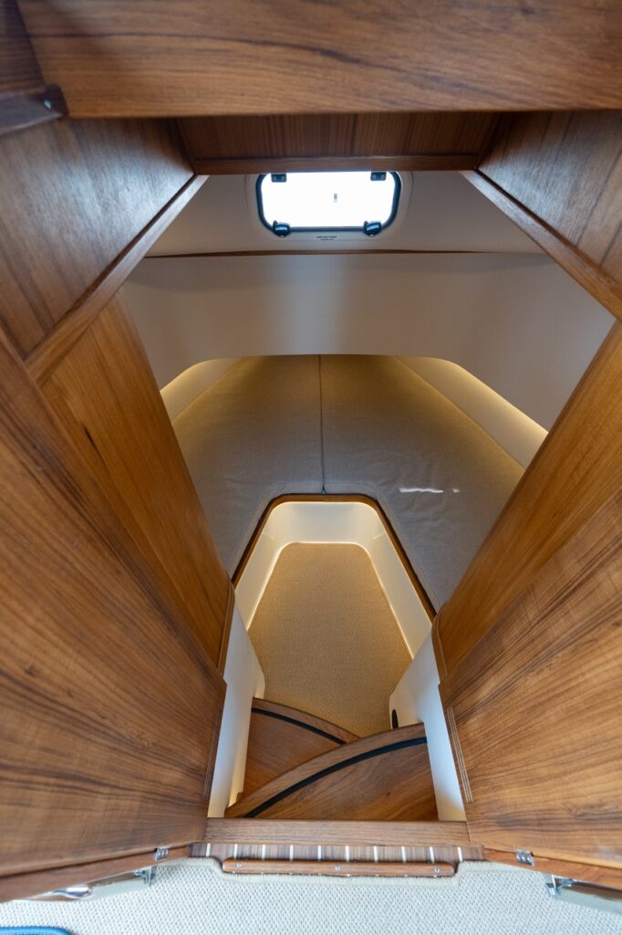 Steps leading down into the split V-berth in the forward cabin, showing illuminated stair treads, teak joinery, twin berth cushions, and a small overhead portlight.