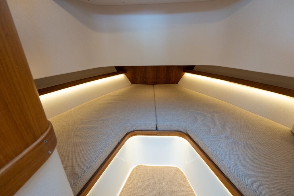 Forward V-berth in the Targa 32 cabin, showing twin berth cushions, indirect LED accent lighting along the hull sides, teak trim, and illuminated step access between the berths.