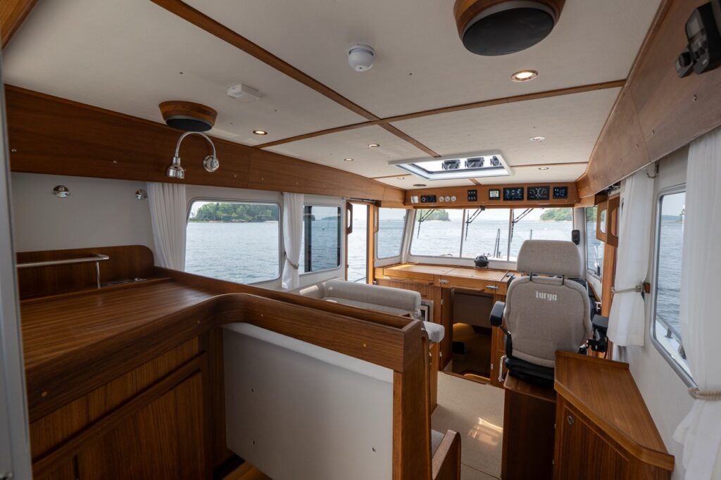 Interior view of the Targa 32 pilothouse and saloon looking forward, showing teak cabinetry, helm seat, wraparound windows, skylight, and seating area with water visible outside.