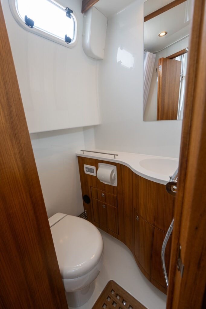 Enclosed marine head on a Targa 32 with toilet, curved vanity with integrated sink, teak cabinetry, mirror, and opening portlight for light and ventilation.