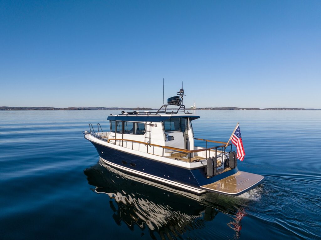 Aerial rear three-quarter view of a Targa 32 cruising on calm blue water in daylight, with an American flag flying aft and the pilothouse and teak decks clearly visible.