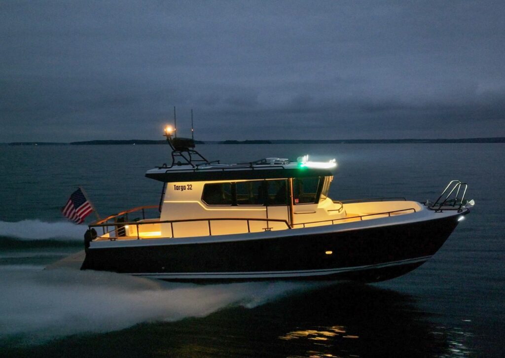 Targa 32 underway at dusk with navigation and deck lights illuminated, cruising across calm water with the pilothouse glowing warmly against a darkening sky.
