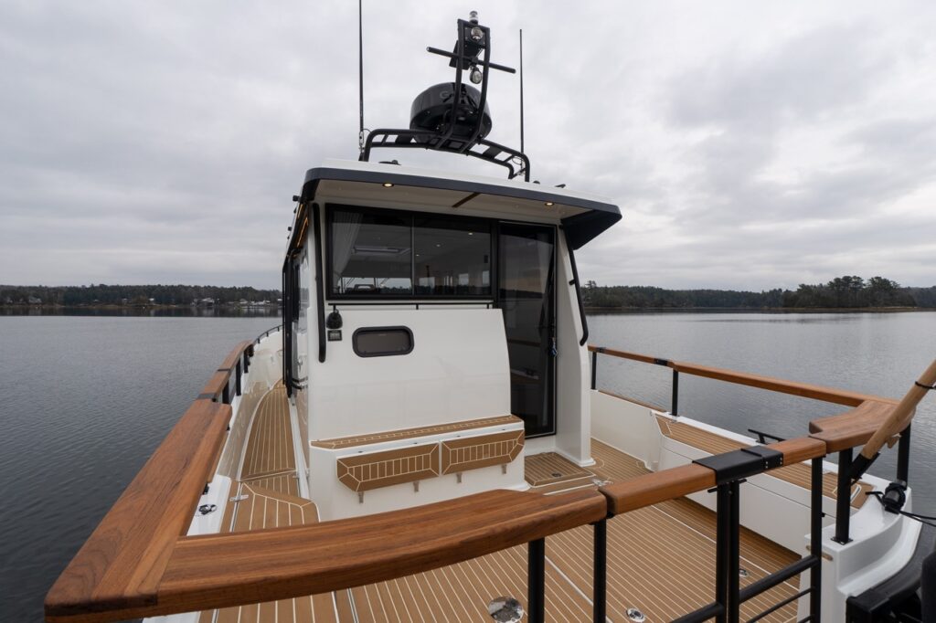 Aft cockpit view of the Targa 32 showing the pilothouse with a hinged aft door swung open, teak-capped cockpit railings, synthetic teak decking, an aft-facing bench with storage, and calm water surrounding the boat.