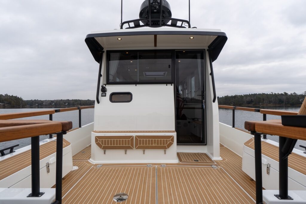 Aft exterior view of the Targa 32 showing the pilothouse entry with a hinged cabin door swung open, a wide aft window into the pilothouse, an overhanging roof with recessed lights, and teak-capped cockpit railings with calm water beyond.