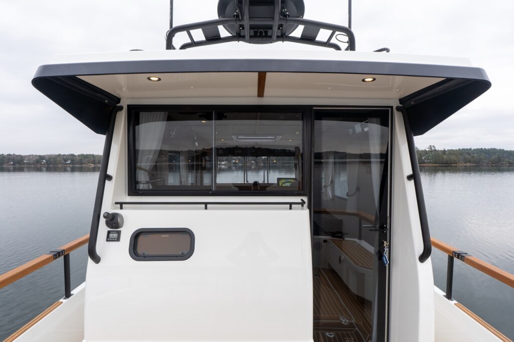Aft exterior view of the Targa 32 showing the pilothouse entry with a hinged cabin door swung open, a wide aft window into the pilothouse, an overhanging roof with recessed lights, and teak-capped cockpit railings with calm water beyond.