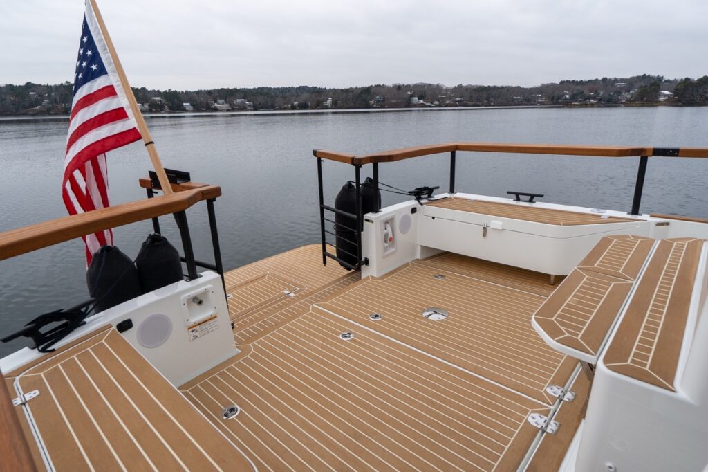 Aft cockpit of the Targa 32 featuring synthetic teak decking, fold-down transom benches, teak-capped railings, fender storage along the transom, and an American flag at the stern with calm water in the background.