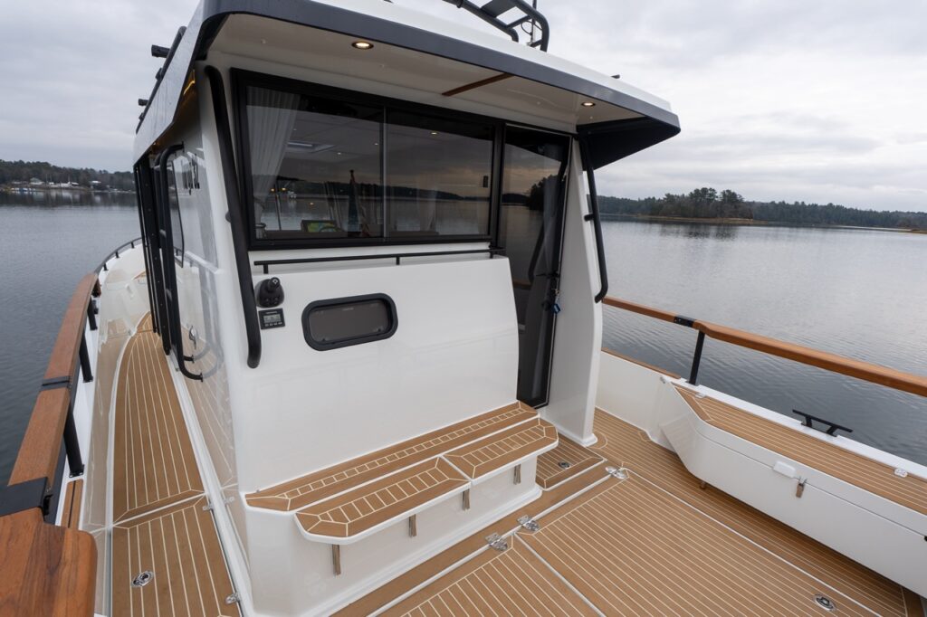 Aft cockpit of a Targa 32 showing the pilothouse rear bulkhead, sliding glass door, integrated steps, teak decking, and wraparound teak handrails with calm water in the background.