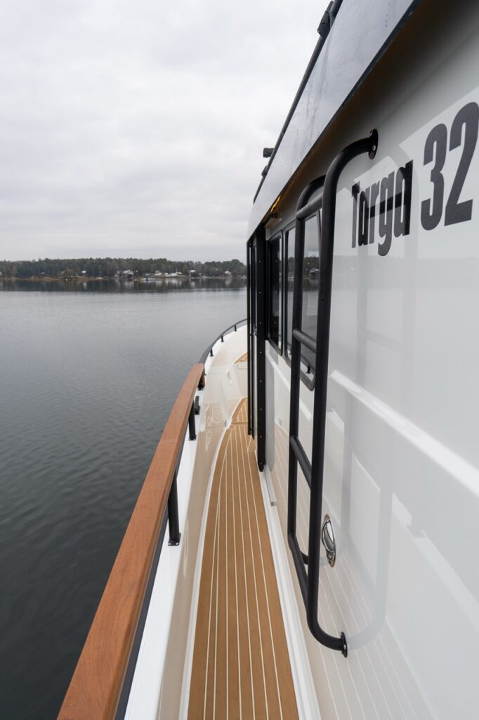 Angled view along the starboard walkaround side deck of a Targa 32, showing teak-patterned decking, a solid teak handrail, black exterior side ladder, pilothouse windows, and “Targa 32” hull-side badging with calm water alongside.