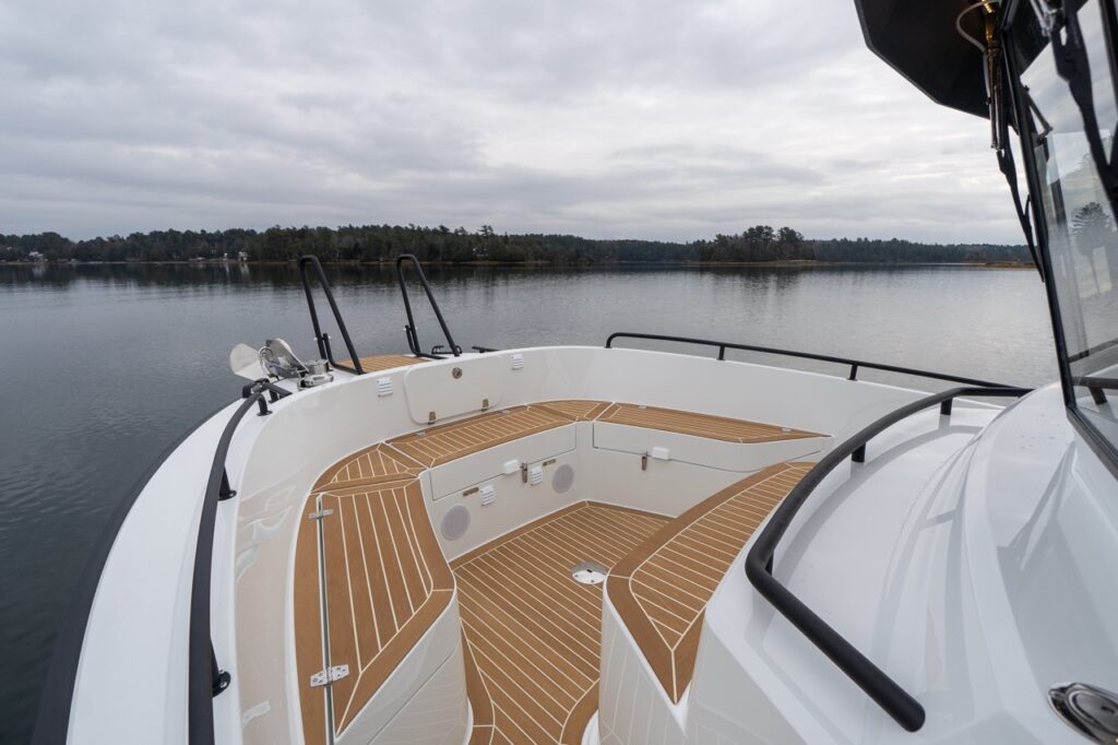 Angled view of the Targa 32 bow seating area from the starboard side, showing wraparound benches with teak-style decking, integrated speakers, anchor windlass, and black safety rails over calm water.