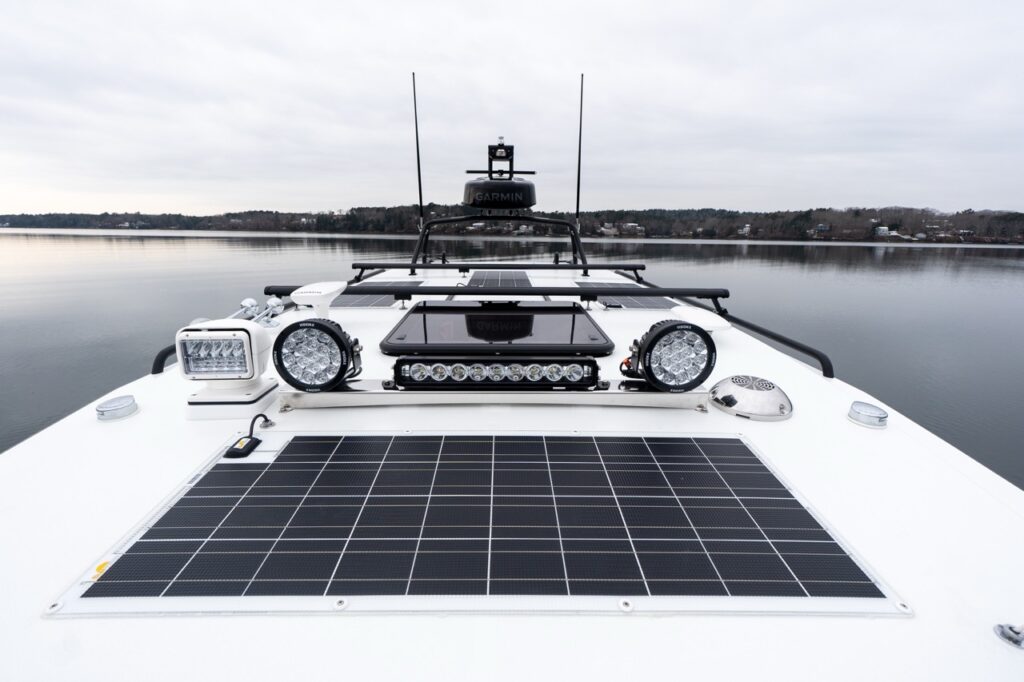 View forward over the Targa 32 pilothouse roof showing integrated solar panels, roof-mounted LED light bar, twin round auxiliary lights, forward searchlight, and Garmin radar array, with calm water and shoreline ahead.