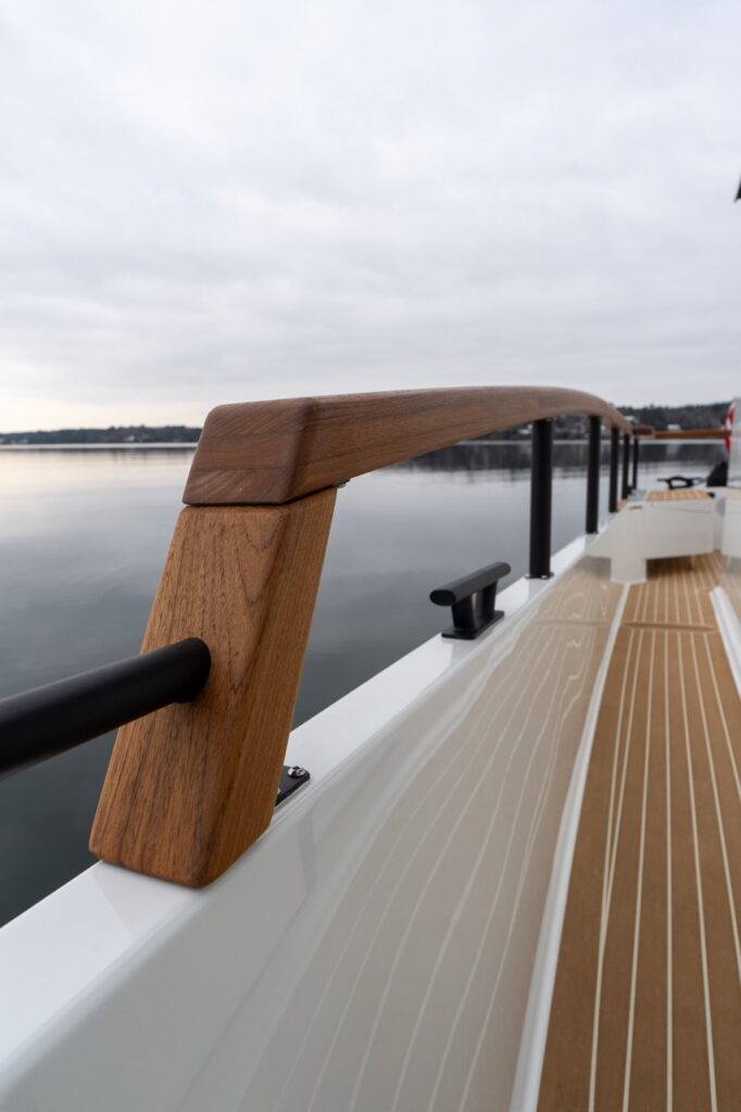 Teak handrails mounted along the walk-around side deck of a Targa, viewed looking aft, with synthetic teak decking underfoot and calm water alongside.