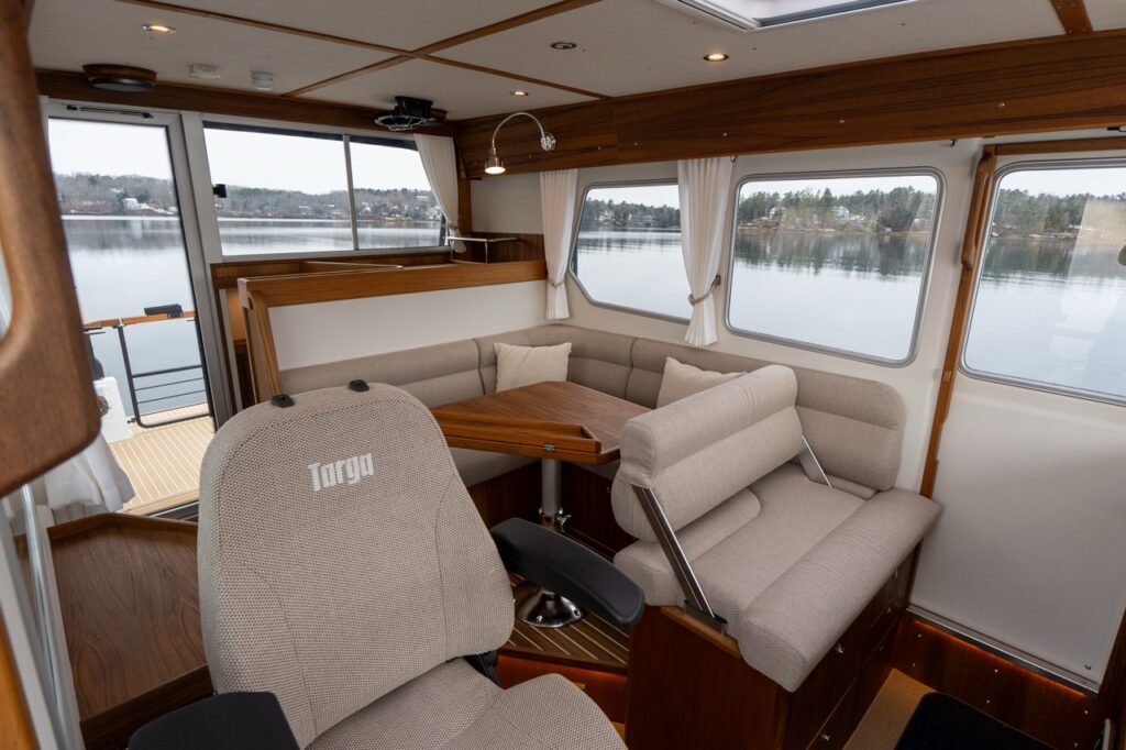 Aft section of the Targa pilothouse showing the L-shaped dinette with upholstered seating and teak table, adjacent to the forward-facing helm seat, with large side windows and an aft door providing expansive water views.