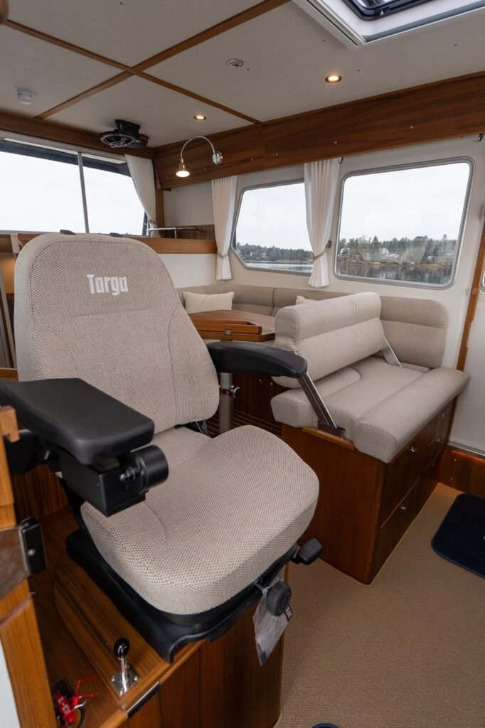 Interior view of the Targa pilothouse showing the upholstered helm seat in the foreground with adjustable armrests, and the aft dinette seating area with wraparound cushions, teak table, and large panoramic windows.