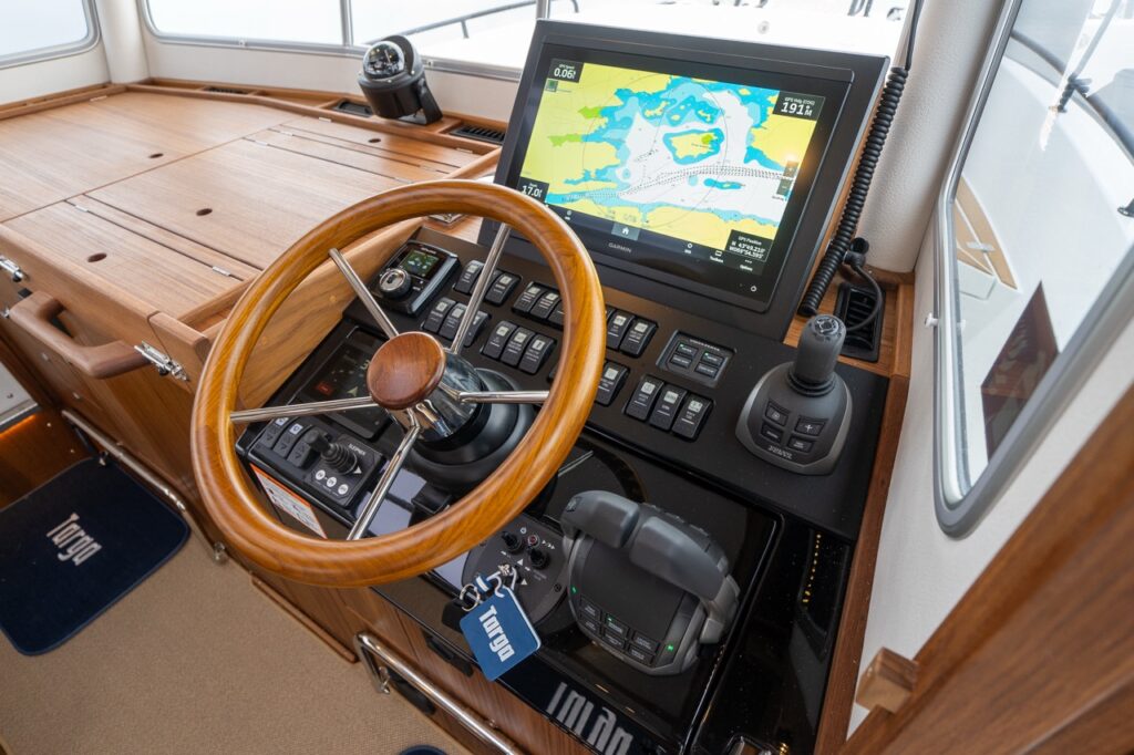 Angled overhead view of the Targa helm featuring a varnished teak steering wheel, Garmin Multi Function Display (MFD) with chartplotter, joystick control, engine throttles, autopilot, and integrated switch panel set into a high-gloss black and teak console.