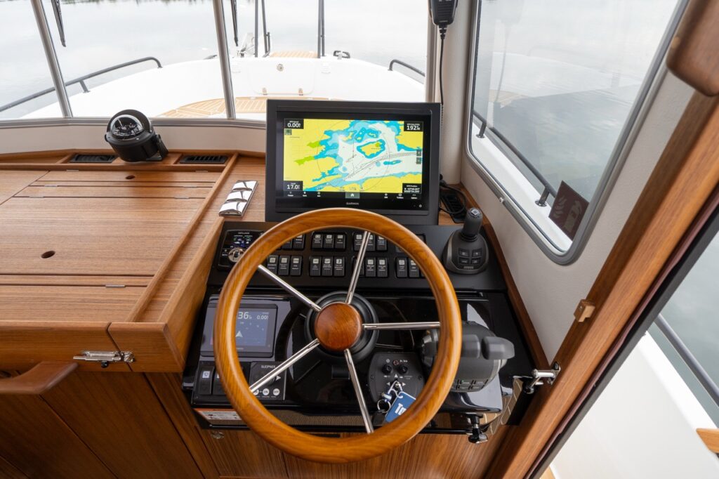 Top-down view of the Targa helm showing a teak-rimmed steering wheel, Garmin Multi Function Display (MFD) with chartplotter data, engine controls, joystick control, compass, and a neatly arranged switch panel within a teak-finished console.