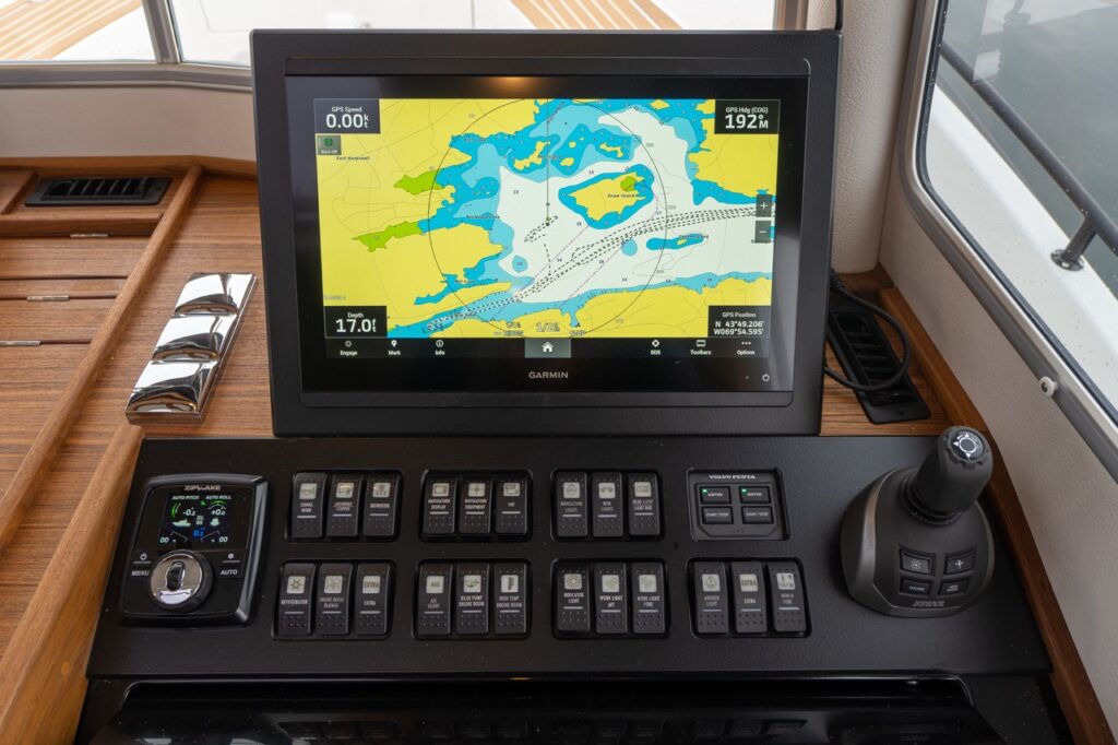 Close-up of the helm console showing a Garmin Multi Function Display (MFD) with nautical chart, depth, speed, and heading data, surrounded by teak trim, integrated joystick control, autopilot interface, and a row of labeled rocker switches below the screen.