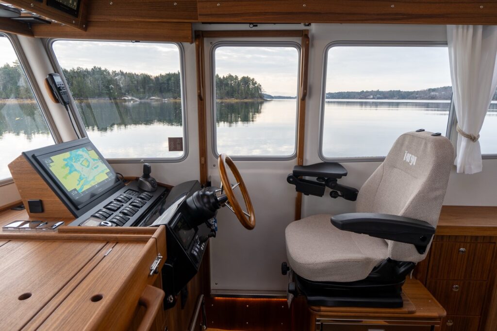 Helm seating area inside the Targa pilothouse showing an adjustable Targa-branded helm chair with armrests, teak helm console with Multi Function Display (MFD), engine controls, wood-rim steering wheel, and large surrounding windows providing clear views over calm water and shoreline.