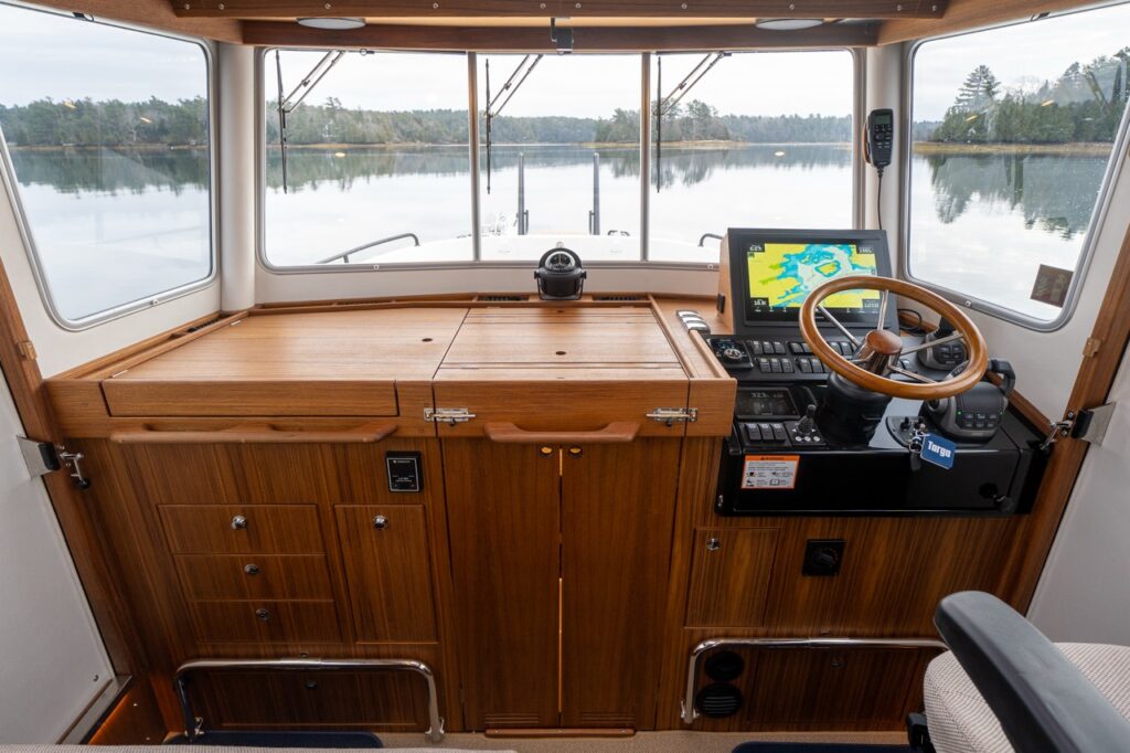 Straight-on view of the Targa 32 pilothouse helm showing the teak helm console with fold-down work surface, integrated storage drawers and cabinets, wood-rim steering wheel, Multi Function Display (MFD), engine controls, compass, and wide forward windows overlooking calm water and shoreline.