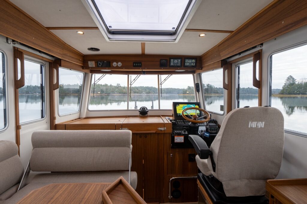 Wide interior view of the Targa 32 pilothouse showing the helm station to starboard with wood-rim steering wheel and navigation display, forward-facing companion seat, wraparound windows, teak cabinetry, and an overhead skylight providing natural light and excellent visibility.