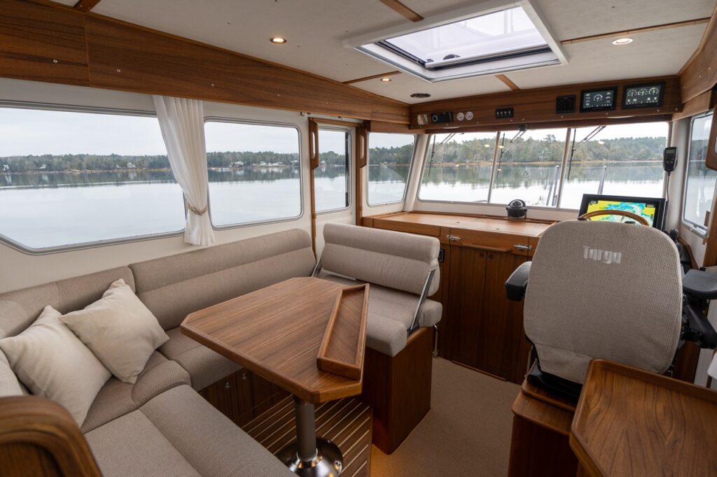 Interior view of the Targa 32 pilothouse showing the L-shaped dinette with teak table to port, forward-facing companion seat, teak helm console to starboard, panoramic windows, and an overhead skylight providing natural light and clear visibility.