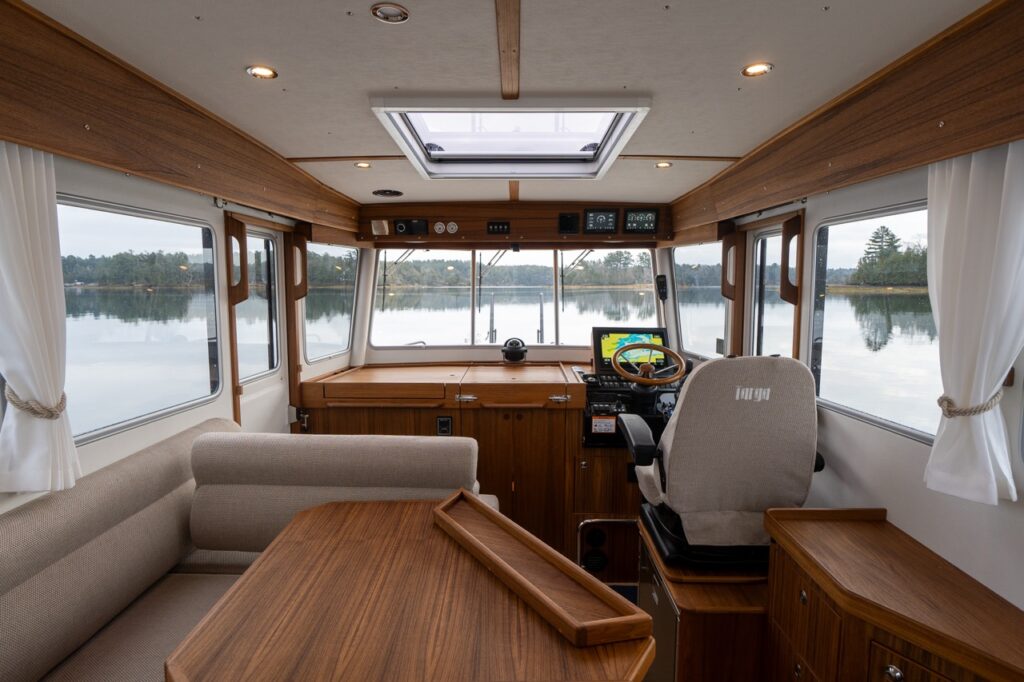 Wide interior view of the Targa 32 pilothouse looking forward, showing the helm station to starboard, forward-facing windows, teak dash and cabinetry, L-shaped dinette with teak table to port, and a large overhead skylight providing natural light.