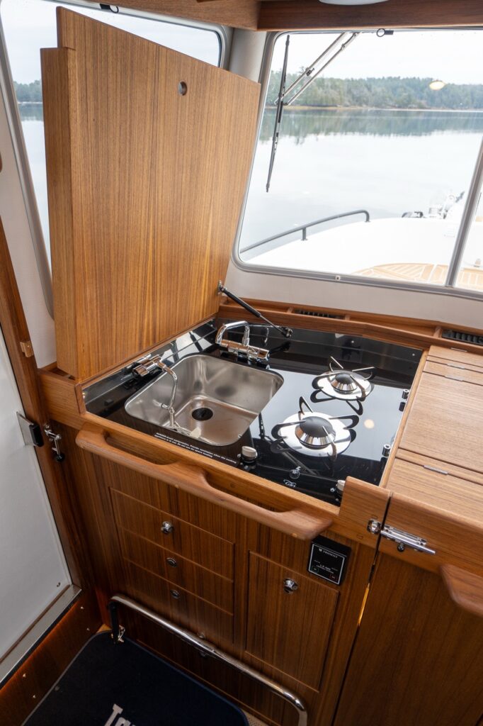 Close-up of the Targa 32 pilothouse galley showing a stainless-steel sink, two-burner cooktop set into a black glass surface, and a hinged teak cover folded upright beneath the forward window, with teak cabinetry below.