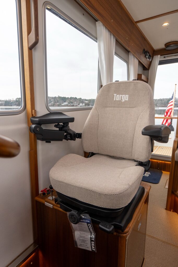 Close-up of the Targa 32 helm seat upholstered in light fabric with “Targa” embroidered on the backrest, featuring adjustable armrests and positioned beside large pilothouse windows.