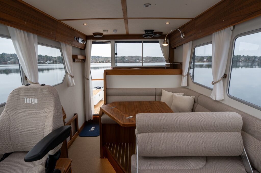 Wide view of the Targa 32 pilothouse interior showing the helm seat, U-shaped dinette with teak table, large windows with curtains, and aft sliding door opening to the cockpit.
