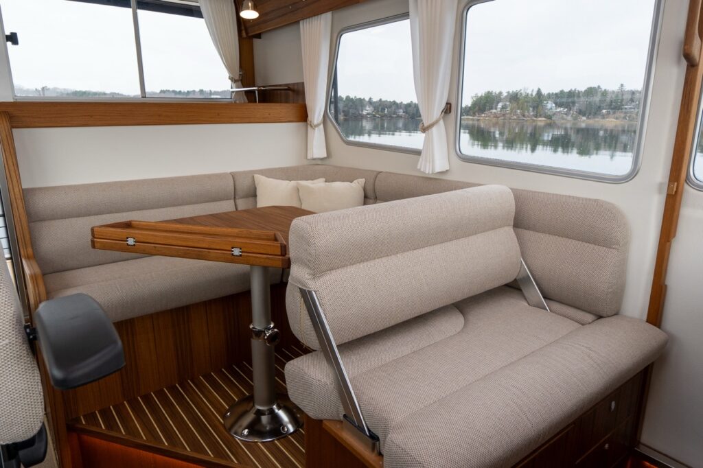 Pilothouse dinette aboard the Targa 32 showing a convertible seating arrangement with a fold-down backrest, teak table, and large side windows overlooking calm water.