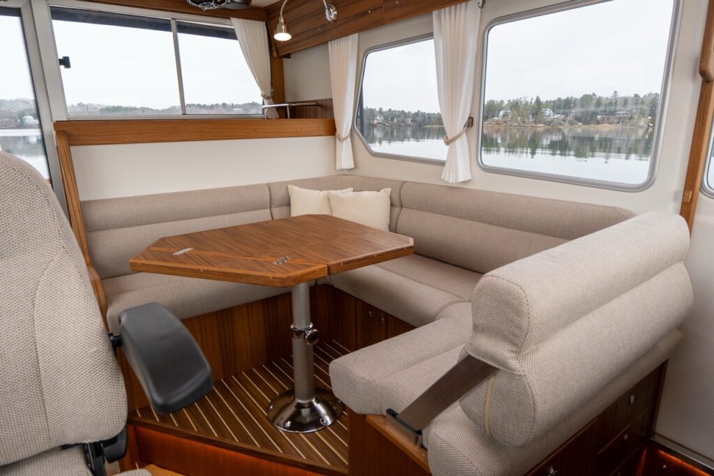 Interior view of the Targa 32 pilothouse dinette featuring a U-shaped upholstered seating area with a teak table, positioned beside large side windows with water views.