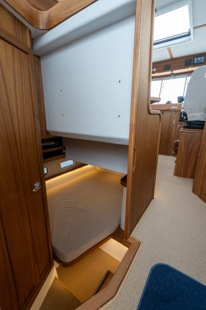 Mid-cabin bunk berth on the Targa 32, showing a compact single sleeping space beneath the helm walkway with upholstered mattress, teak cabinetry, hull-side portlight, and soft LED floor lighting.