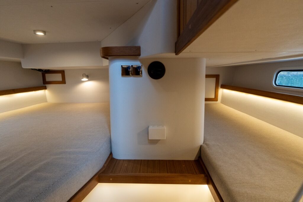 Aft cabin on the Targa 32 showing both the double berth to port and the single twin berth to starboard, separated by a central structural column, with upholstered mattresses, hull-side portlights, teak trim, and warm LED accent lighting.